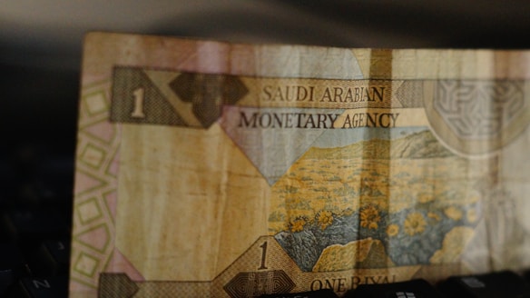 A close-up view of a paper currency note from Saudi Arabia, featuring floral imagery and intricate patterns. The note has the denomination of one rial, with the text 'Saudi Arabian Monetary Agency' printed on it.