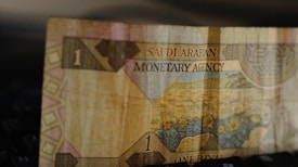 A close-up view of a paper currency note from Saudi Arabia, featuring floral imagery and intricate patterns. The note has the denomination of one rial, with the text 'Saudi Arabian Monetary Agency' printed on it.