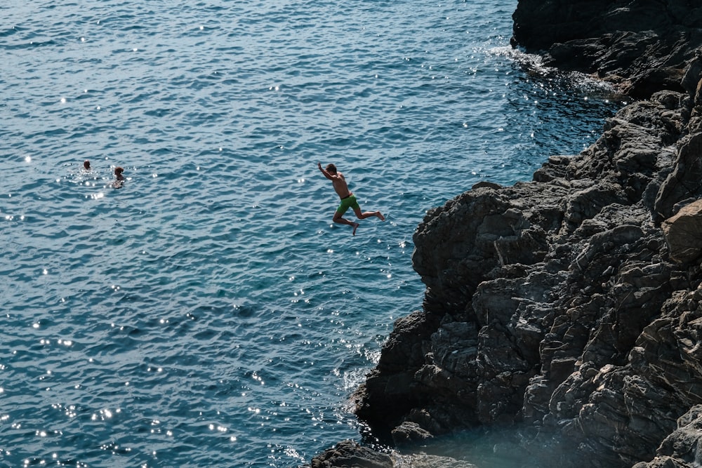 man in green shorts jumping on rocky cliff by the sea during daytime