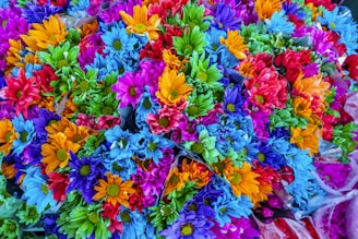 A vibrant bouquet of artificial flowers showcasing a variety of colors including blue, green, orange, purple, and red. Each flower displays a full bloom, creating a dense and colorful arrangement.