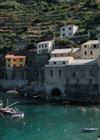 A coastal landscape features several multi-story buildings with a rustic appearance situated on a terraced hillside. Below, the calm turquoise waters of a bay are dotted with small boats and people swimming. The scene is bathed in natural light, highlighting the lush greenery on the hillside.