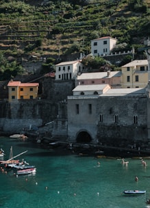 A coastal landscape features several multi-story buildings with a rustic appearance situated on a terraced hillside. Below, the calm turquoise waters of a bay are dotted with small boats and people swimming. The scene is bathed in natural light, highlighting the lush greenery on the hillside.