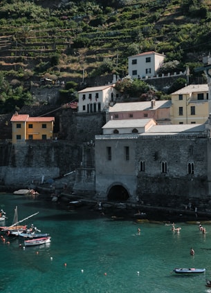 A coastal landscape features several multi-story buildings with a rustic appearance situated on a terraced hillside. Below, the calm turquoise waters of a bay are dotted with small boats and people swimming. The scene is bathed in natural light, highlighting the lush greenery on the hillside.
