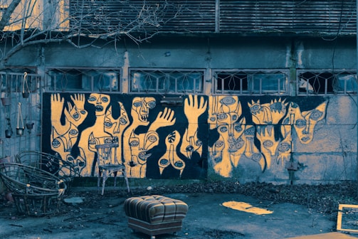 A wall covered with abstract graffiti art depicting various faces and hands with exaggerated features. The artwork employs a palette of black and yellow, creating a striking contrast. Below the graffiti, there are several chairs, one of which is overturned. The surrounding environment appears neglected, with bare branches from a tree and scattered leaves on the ground, indicating an urban setting.