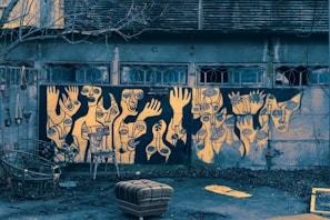 A wall covered with abstract graffiti art depicting various faces and hands with exaggerated features. The artwork employs a palette of black and yellow, creating a striking contrast. Below the graffiti, there are several chairs, one of which is overturned. The surrounding environment appears neglected, with bare branches from a tree and scattered leaves on the ground, indicating an urban setting.