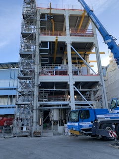 A multi-story industrial building under construction, featuring scaffolding on the left side and a blue crane on the right. The structure has exposed steel beams and various pieces of construction equipment. The ground level shows a large metal tank, and the site is surrounded by safety barriers.