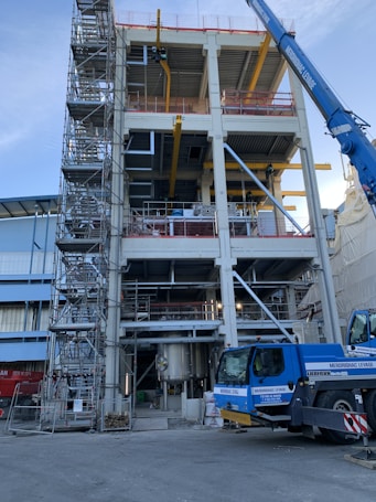 A multi-story industrial building under construction, featuring scaffolding on the left side and a blue crane on the right. The structure has exposed steel beams and various pieces of construction equipment. The ground level shows a large metal tank, and the site is surrounded by safety barriers.