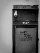 A black and white photo of a door labeled '310C' with the text 'Evans & Peel Detective Agency' on a glass panel. There is a security camera above the door and a textured, aged appearance to the wood and glass.