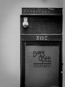 A black and white photo of a door labeled '310C' with the text 'Evans & Peel Detective Agency' on a glass panel. There is a security camera above the door and a textured, aged appearance to the wood and glass.
