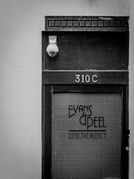 A black and white photo of a door labeled '310C' with the text 'Evans & Peel Detective Agency' on a glass panel. There is a security camera above the door and a textured, aged appearance to the wood and glass.