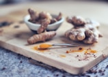 A wooden board with fresh turmeric roots and a small spoon of ground turmeric. In the background, a ceramic bowl contains additional turmeric roots, slightly out of focus.
