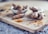 A wooden board with fresh turmeric roots and a small spoon of ground turmeric. In the background, a ceramic bowl contains additional turmeric roots, slightly out of focus.