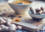 A close-up view of a wooden cutting board with fresh turmeric roots and a spoon of ground turmeric. A small white bowl holds more turmeric roots, and a blurred soup bowl is visible in the background. The setting has an earthy and natural feel, with scattered pieces of spices adding detail.