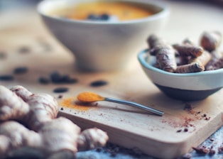 A close-up view of a wooden cutting board with fresh turmeric roots and a spoon of ground turmeric. A small white bowl holds more turmeric roots, and a blurred soup bowl is visible in the background. The setting has an earthy and natural feel, with scattered pieces of spices adding detail.