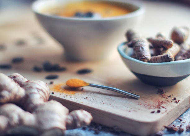 A close-up view of a wooden cutting board with fresh turmeric roots and a spoon of ground turmeric. A small white bowl holds more turmeric roots, and a blurred soup bowl is visible in the background. The setting has an earthy and natural feel, with scattered pieces of spices adding detail.