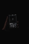A dark, moody image showing a hand holding a smartphone with a music player app open. The screen displays a song titled 'PAID MY DUES' by NF. The background is completely black, emphasizing the illuminated screen.