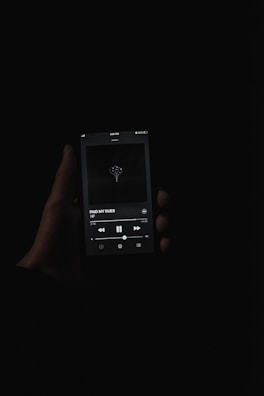 A dark, moody image showing a hand holding a smartphone with a music player app open. The screen displays a song titled 'PAID MY DUES' by NF. The background is completely black, emphasizing the illuminated screen.