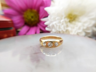 A gold ring with three diamond stones set in it is placed on a reflective surface. The background features two blurred flowers, one pink and one white, adding a soft, elegant touch to the composition.