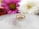A gold ring with three diamond stones set in it is placed on a reflective surface. The background features two blurred flowers, one pink and one white, adding a soft, elegant touch to the composition.