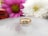 A gold ring with three diamond stones set in it is placed on a reflective surface. The background features two blurred flowers, one pink and one white, adding a soft, elegant touch to the composition.