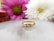 A gold ring with three diamond stones set in it is placed on a reflective surface. The background features two blurred flowers, one pink and one white, adding a soft, elegant touch to the composition.