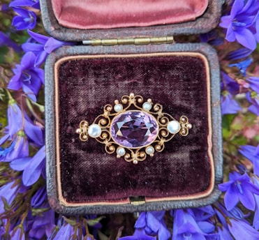 An ornate brooch featuring a large, faceted purple gemstone at the center, surrounded by a delicate arrangement of smaller pearls set in intricate gold metalwork. It is placed inside an open vintage jewelry box lined with dark velvet and pale pink satin. The background is filled with vibrant purple flowers, adding a rich and luxurious feel to the composition.