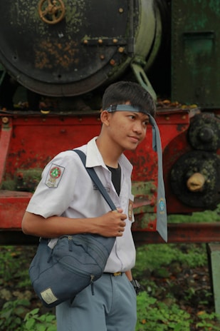 A young person in a school uniform stands in front of an old, weathered locomotive. They have a tie draped over their forehead and are holding a blue satchel. The locomotive behind them is rusted with green and red colors, adding a vintage aesthetic to the scene.