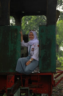 A person is wearing a white hijab and a light blue uniform, sitting inside an old train carriage with open doors. The background includes green foliage visible through the open sections of the train.
