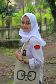 A person wearing a white hijab, school uniform, and a watch smiles while holding a beige tote bag with a playful graphic of glasses and eyes. The background features a lush, green natural setting with trees and a pathway covered in fallen leaves.