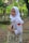 A person wearing a white hijab, school uniform, and a watch smiles while holding a beige tote bag with a playful graphic of glasses and eyes. The background features a lush, green natural setting with trees and a pathway covered in fallen leaves.