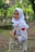 A person wearing a white hijab, school uniform, and a watch smiles while holding a beige tote bag with a playful graphic of glasses and eyes. The background features a lush, green natural setting with trees and a pathway covered in fallen leaves.