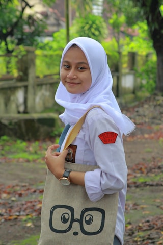 A person wearing a white hijab, school uniform, and a watch smiles while holding a beige tote bag with a playful graphic of glasses and eyes. The background features a lush, green natural setting with trees and a pathway covered in fallen leaves.