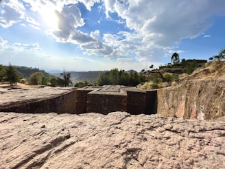 An ancient rock-hewn church is carved into the ground, surrounded by rugged terrain and scattered vegetation. The clear blue sky with scattered clouds is illuminated by bright sunlight, casting shadows and adding to the serene atmosphere. The background features hills with sparse trees and greenery further enhancing the natural beauty. The detailed carvings on the church's roof can be seen, indicative of intricate craftsmanship.
