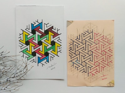 Two geometric art designs placed side by side on paper. The left design features a colorful pattern with interconnected shapes in blue, green, yellow, pink, and black. The right design is made up of complex interlocking lines in black and red on a beige background. A dried branch lies on the lower left corner, adding a natural element to the composition.