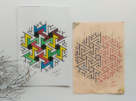 Two geometric art designs placed side by side on paper. The left design features a colorful pattern with interconnected shapes in blue, green, yellow, pink, and black. The right design is made up of complex interlocking lines in black and red on a beige background. A dried branch lies on the lower left corner, adding a natural element to the composition.