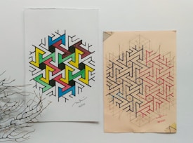 Two geometric art designs placed side by side on paper. The left design features a colorful pattern with interconnected shapes in blue, green, yellow, pink, and black. The right design is made up of complex interlocking lines in black and red on a beige background. A dried branch lies on the lower left corner, adding a natural element to the composition.