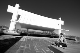 A person is sitting cross-legged in a meditative pose on a tiled surface next to a large, modern architectural structure with clean, sweeping lines and a prominent overhang. The sky is clear, and there's a railing behind the person suggesting an elevated viewpoint.
