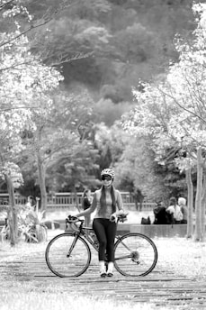 A person wearing cycling gear including a helmet and sunglasses stands holding a road bicycle. They are positioned on a path lined with trees. In the background, a group of people sit together, possibly socializing or resting. The scene is set in a natural environment with a hilly or forested area in the distance.