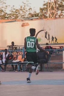 A person in a green basketball jersey with the number 00 and the name A. Azhary on the back is playing on an outdoor basketball court. The court has a few spectators seated on benches in the background. The lighting suggests late afternoon or early evening with shadows extending across the ground. The surroundings include trees and a mural painted on a nearby wall.