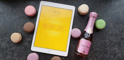 A white tablet with a yellow screen displaying the message 'No Bad Days' placed on a dark surface, surrounded by colorful macarons including pink, brown, green, and beige. Next to the tablet, there is a small bottle of pink sparkling wine.