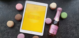 A white tablet with a yellow screen displaying the message 'No Bad Days' placed on a dark surface, surrounded by colorful macarons including pink, brown, green, and beige. Next to the tablet, there is a small bottle of pink sparkling wine.