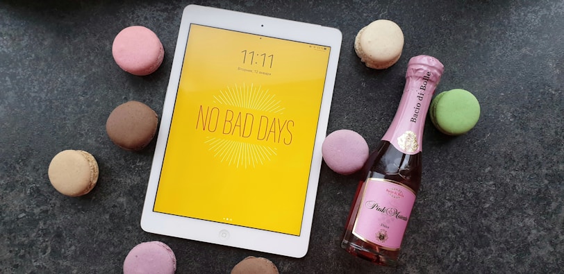 A white tablet with a yellow screen displaying the message 'No Bad Days' placed on a dark surface, surrounded by colorful macarons including pink, brown, green, and beige. Next to the tablet, there is a small bottle of pink sparkling wine.