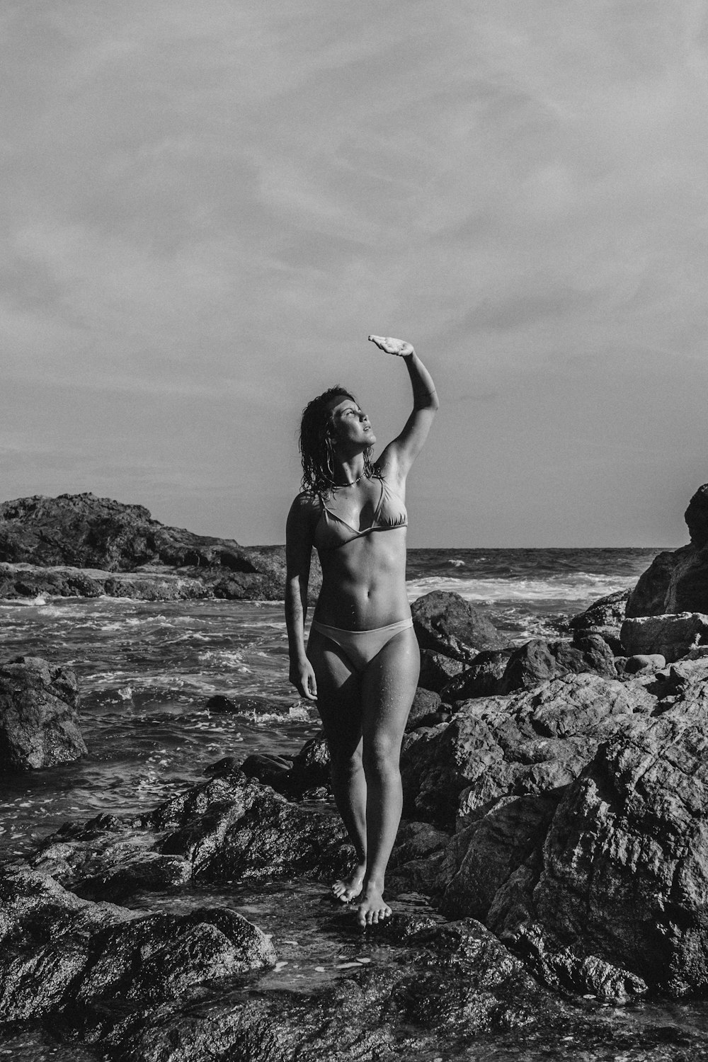 grayscale photo of woman in bikini standing on rock near sea