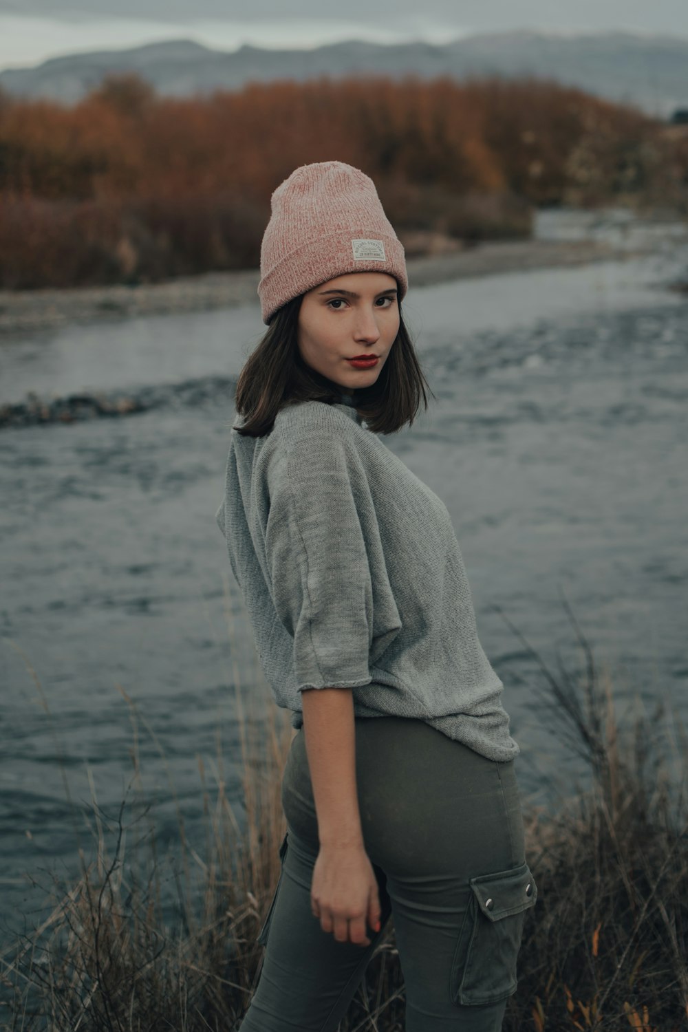 woman in gray sweater and pink knit cap standing near body of water during daytime