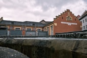 A historic brick building with an ornate gable façade and a sign labeled 'STÄDTISCHES ELEKTRIZITÄTSWERK' alongside other industrial structures. The scene appears to be part of a small power plant or utility station, bounded by metal fencing and situated near a body of water or channel.