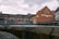 A historic brick building with an ornate gable façade and a sign labeled 'STÄDTISCHES ELEKTRIZITÄTSWERK' alongside other industrial structures. The scene appears to be part of a small power plant or utility station, bounded by metal fencing and situated near a body of water or channel.