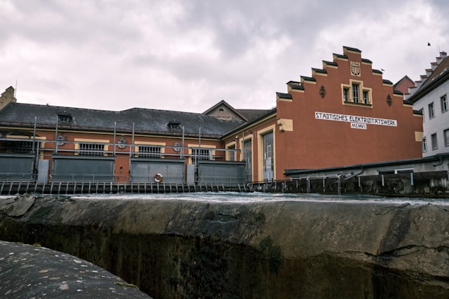 A historic brick building with an ornate gable façade and a sign labeled 'STÄDTISCHES ELEKTRIZITÄTSWERK' alongside other industrial structures. The scene appears to be part of a small power plant or utility station, bounded by metal fencing and situated near a body of water or channel.