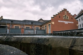 A historic brick building with an ornate gable façade and a sign labeled 'STÄDTISCHES ELEKTRIZITÄTSWERK' alongside other industrial structures. The scene appears to be part of a small power plant or utility station, bounded by metal fencing and situated near a body of water or channel.