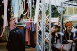 A bustling outdoor market scene with several people browsing clothes on display racks. Brightly colored garments hang alongside streamers. A canopy provides shade, while a sign reading 'PALETAS' is visible in the background. Shoppers are engaged in conversation and appear to be enjoying the lively atmosphere.