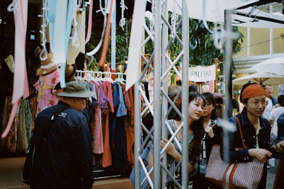 A bustling outdoor market scene with several people browsing clothes on display racks. Brightly colored garments hang alongside streamers. A canopy provides shade, while a sign reading 'PALETAS' is visible in the background. Shoppers are engaged in conversation and appear to be enjoying the lively atmosphere.